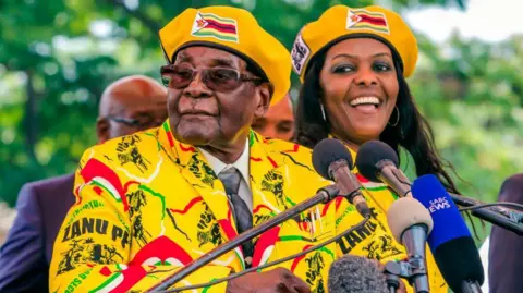 AFP via Getty Images Grace and Robert Mugabe, dressed in bright yellow Zanu-PF clothes and berets stand next to each other at some microphones. 